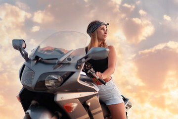 Young woman poses sitting behind the wheel of a motorcycle. Golden sunset and motorbike on the...
