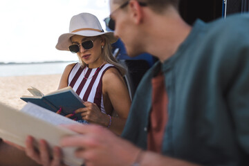 Young couple sitting together in front of van, camping and reading book.