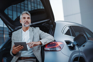 Businessman using tablet while charging car at electric vehicle charging station, closeup.