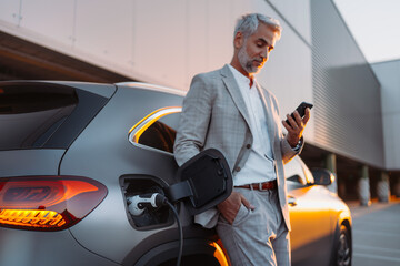 Businessman holding smartphone while charging car at electric vehicle charging station, closeup.
