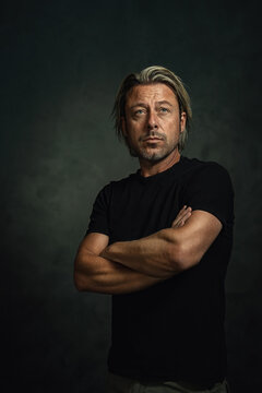 Serious blond man with medium length hair and a stubble beard in a black t-shirt stands with crossed arms in front of a dark gray wall.