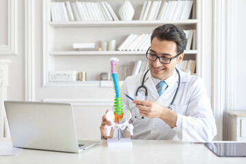 Young handsome male doctor orthopedist demonstrating the problem on spine bone model on the desk in his workplace - obrazy, fototapety, plakaty