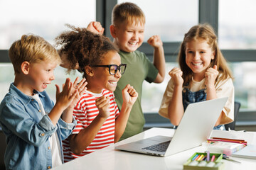 Cheerful  schoolkids   looking at laptop screen and celebrate   successful completion of collective school work   during online lesson