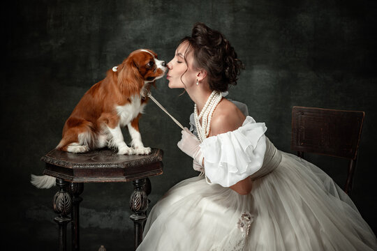 Kiss. Vintage portrait of young elegant woman in image of medieval person in renaissance style dress with little cute dog isolated on dark background.