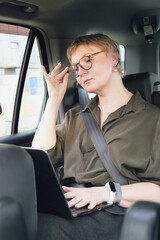 Business woman in glasses is sitting in the back seat of a car. Business woman is working on laptop in car. Serious middle aged woman with glasses