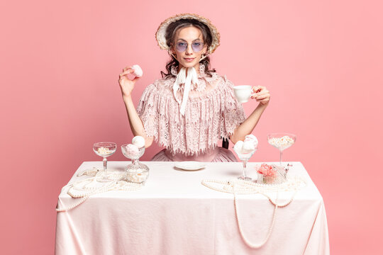 Morning coffee. Young pretty woman with retro style hairdo in bonnet and vintage dress sitting at holiday table isolated on pink background.