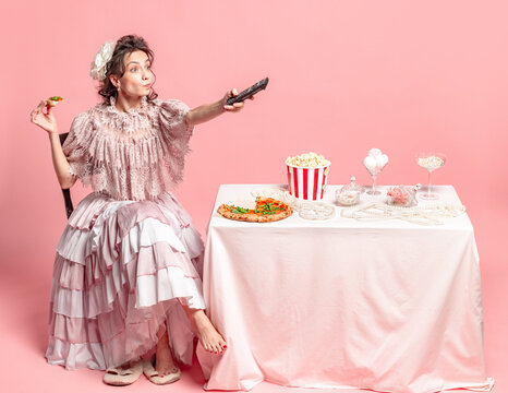 Cozy evening. Young beautiful woman with retro style hairdo wearing medieval dress sitting at served table isolated on pink background.