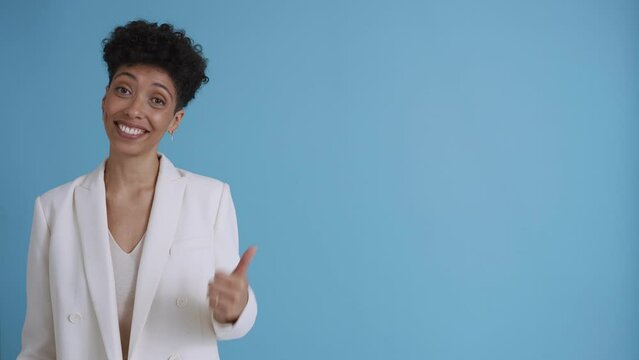 Happy hispanic curly haired woman wearing white jacket talking and pointing to the copyspace in the blue studio