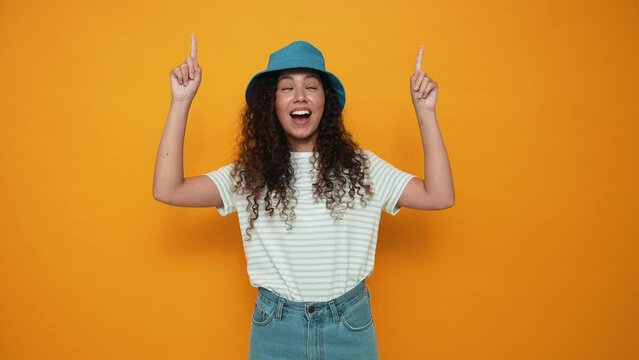 Happy brunette curly haired woman wearing panama hat pointing fingers upwards in the yellow studio