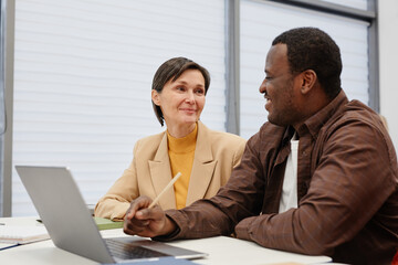 Mature woman talking to African teacher while they sitting together at desk and learning computer program on laptop