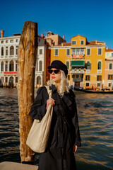 Elegant confident woman wearing trendy baker boy cap, cat eye sunglasses, classic blue trench coat, with big shoulder hobo bag, posing in street of Venice. Fashion, travel, lifestyle conception