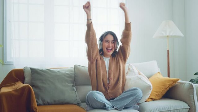 Happy young asian woman wear wireless headphones having fun listen music in smartphone and sit on sofa in living room, Female enjoys listening chill music audio sound feeling no stress at home.