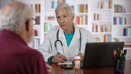 Mature senior female doctor talking to male patient in modern clinic with library background.