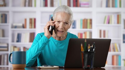 Stressed senior woman on smartphone, phone, with reaction to bad news in front of laptop computer in home office.