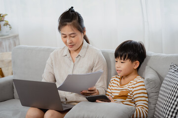 A middle-aged mother sits on the sofa intent on laptop, and her son sits on tablet next to her in the living room. The boy tried to pull the paper out of mother's hand, wanting to interest in him