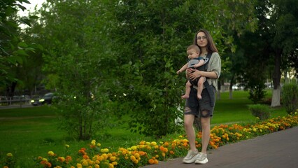 A young woman walks along the alley in the park with her son. The girl in glasses walks. Newborn son in a kangaroo-backpack.
