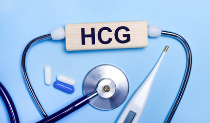On a light gray background, a stethoscope, an electronic thermometer, pills, a wooden block with the text HCG Human Chorionic Gonadotrophin. Medical concept.