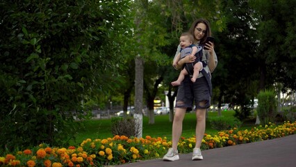 A child in a kangaroo-backpack in his mother's arms. Young woman in glasses is texting on the phone in the park.