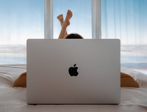 2022, Europe: Unrecognizable girl lying in bed and working behind macbook pro laptop on panoramic windows background. Young woman lying on bed and using mackbook on sea background with blue sky