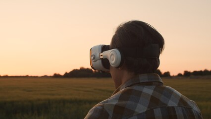 Fototapeta premium Farmer in virtual reality helmet in front of a sunset agricultural landscape. Man in a countryside field. Country life, food production, farming and technology concept