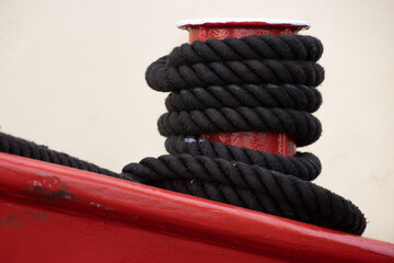 Black rope tied to a red metal mooring bollard - obrazy, fototapety, plakaty