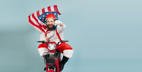 Young adult man in a santa costume in a superman pose with a fluttering usa flag in the form of a cape and a helmet rides a red electric scooter on a blue isolated background with copy space