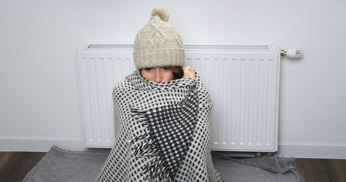 Woman cold in apartment. Young woman in sweater, scarf, hat, warm chalet sitting alone near radiator, try warming indoor. Poor heating in Europe house concept. Shaking, shivering from chilly in room 