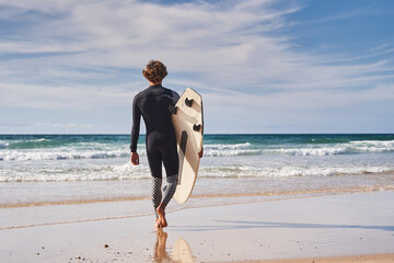 Back view of the young man with surfboard going for surfing in the sea