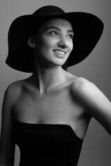 Black and white studio fashion portrait of elegant young and sexy woman in black wide hat and classic evening dress. Stylish elegant woman looking aside of camera in grey background with copy space