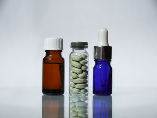 There are a glass jar with green pharmaceutical pills, blue pipette with medical drops and brown medical bottle with oil on the mirror background