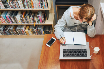 Student learning in university library. Young woman reading textbook and making notes using computer. Focused student studying for college exams. Using technology