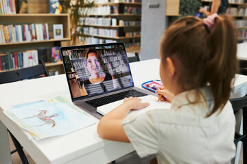 Student litttle girl having video class remotely with her teacher on laptop sitting at desk in...