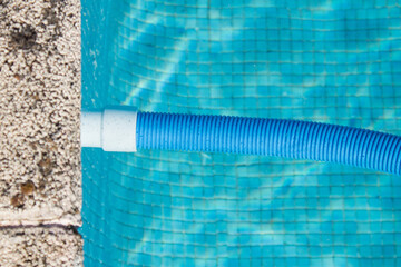 A connected blue pool cleaner hose floats in the pool water next to the stone curb. Pool cleaning and maintenance.