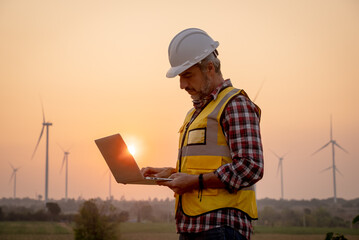Portrait of engineer wearing yellow vest and white helmet using a computer laptop on site at wind...