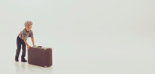 Portrait of little boy, child in checkered shirt pushing vintage suitcase isolated over grey studio background. Travel