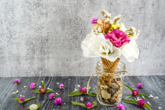 Ice cream cone with a flower bouquet in it. Cone is placed in a small glass jar, tied with twine, and pebbles in it. Gray background. Still life image.
