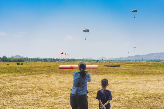 Parents take video clips and photo with smart phone and watch with worry and concern during parachute training from airplane for army cadet with blurred image of parachute and landscape in background.