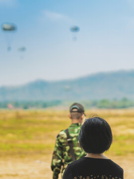 Back view of female parents look with worry and concern during parachute training from airplane for army cadet with blurred image of parachute and landscape in background. Family relationship concept.