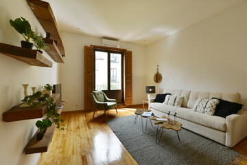 Living room of a house with large beige fabric sofa with cushions, balcony with wooden shutters, raw wooden shelves with plants and original wooden floors