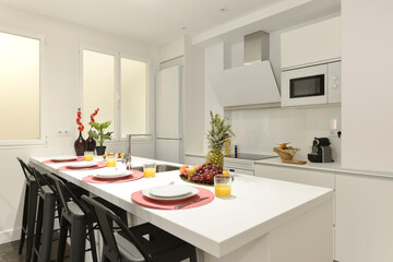 Kitchen with white wooden furniture and integrated matching appliances, island with white wooden countertop, with fruits and juices and industrial style black metal high stools