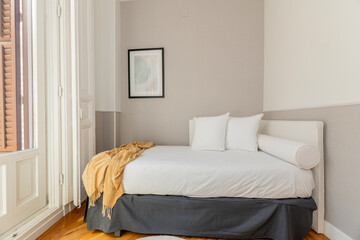 Youthful bedroom with white fabric upholstered headboard, matching divan, white cushions and balcony with white wooden windows and shutters