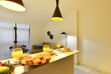Living room with an open kitchen, with a white rectangular dining table with breakfast service with...