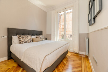 Bedroom with gray fabric upholstered headboard, matching cushions and pillows, large balcony with white windows and shutters, matching blankets, original wooden floor and decorative mirror on the wall