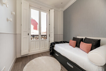 Youth bedroom with headboard upholstered in white fabric, matching cushions and pillows, large balcony with white windows and shutters, matching blankets, light wooden floor