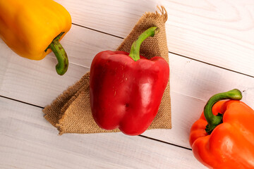 Three whole organic fresh juicy tasty bell peppers, close-up, on a jute napkin lying on a painted white table top made of wood, top view.