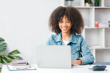 American teenage woman sitting in white office with laptop, she is a student studying online with...