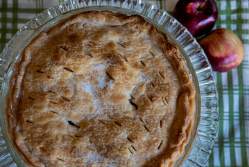 Closeup of beautiful homemade apple pie with sugared topping on glass plate and green and white checkered cloth with apples on side