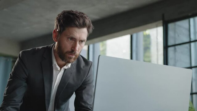 Serious 40s bearded businessman entrepreneur employee standing in office looking at papers report information papers check data in online net service in computer typing working paperwork deadline