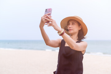 An Asian woman stands to take a selfie with her mobile phone in the sand during a summer vacation on the beach