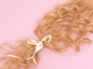 Curled natural long blonde hair tied with a white bow on a pink background. Hair donation. Hair...
